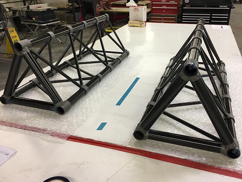 Two marine sensor masts are assembled side-by-side for a dry fit. They are laying on their sides on a white table top on plastic bubble wrap. Table is in a manufacturing environment. 
